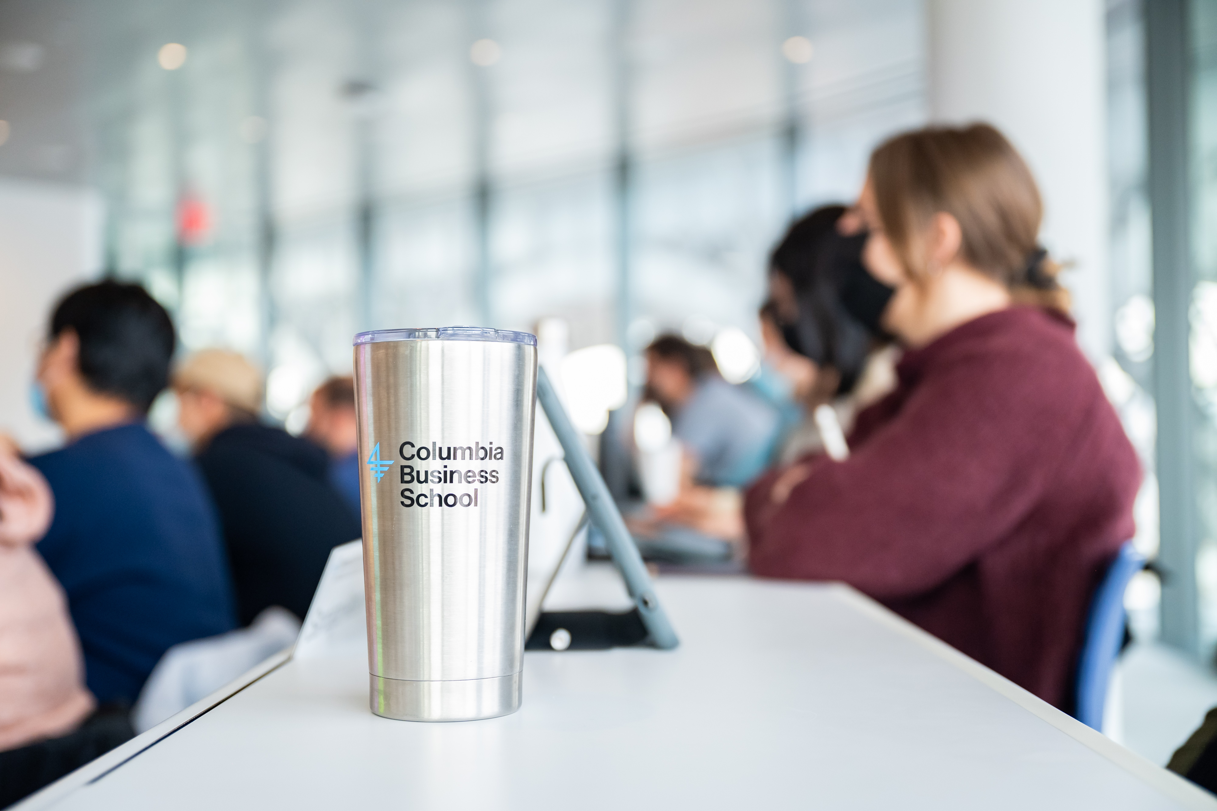 A CBS-branded thermos sits on a desk in a classroom, with rows of students in the background