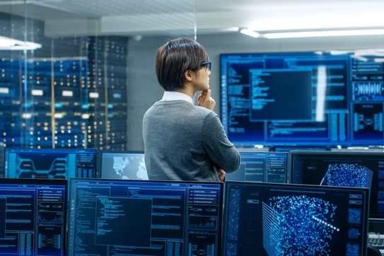 A man in a room full of a computer screens, staring at a screen.