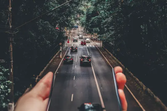 A hand holds out a smartphone; its image of cars driving down the road seems to become the road itself. Photo by Matheus Bertelli.