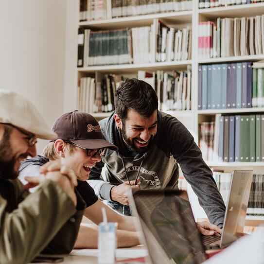 A group of people sitting around a desk with a laptops laughing.